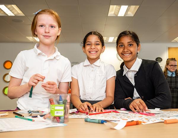 Three children in school uniform stand smiling in front of desk with paper and colouring pens